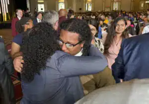 A young man and woman embrace warmly amidst a crowd, highlighting a moment of celebration or connection. The setting appears to be a large event with many attendees in the background.