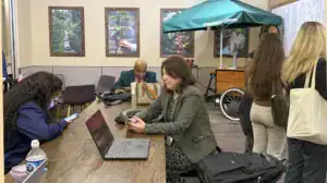 A busy coffee shop scene featuring a woman using her phone at a table with a laptop, while a man sits in the background with a bag. Other people are chatting nearby with a Starbucks cart in view.