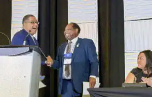 Two men shake hands and smile at a podium during a conference, with a woman seated at a table nearby. The atmosphere appears friendly and engaging.