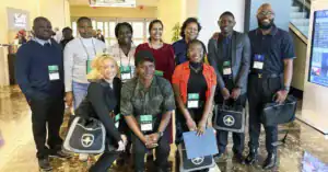 A diverse group of people stands in a lobby, smiling and posing for a photo. They are wearing name tags and carrying bags, with a large digital display visible in the background.