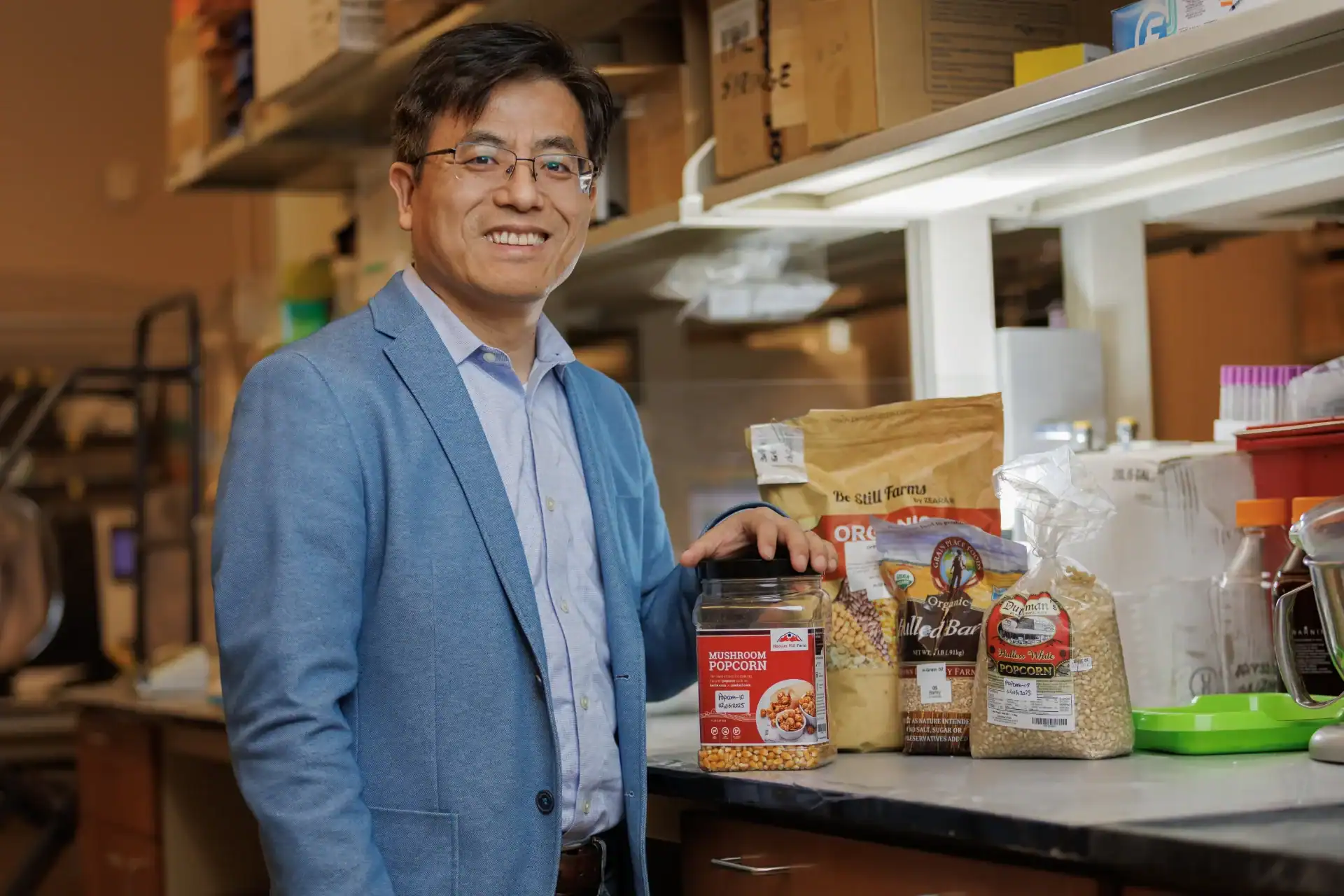 A smiling man in a blue blazer stands in a lab setting, holding a jar of mushroom popcorn. Several bags of grains and ingredients are displayed on the counter behind him.