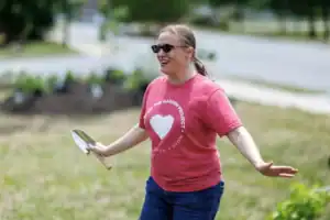 A woman in a red shirt smiles and gestures while holding a gardening tool, surrounded by plants in a grassy area on a sunny day.
