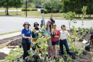 A group of six people smiles and poses in a community garden, each holding plants. They stand among newly planted trees and greenery, showcasing a vibrant outdoor scene on a sunny day.