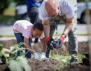 A child and an adult are planting in a garden. The child, in a pink shirt, is focused on digging, while the adult, wearing gloves, is holding a small potted plant. They work together among rows of soil and plants.