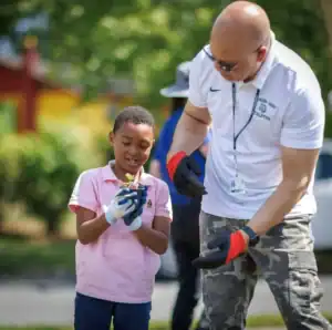 A young boy in a pink shirt holds a small plant while smiling at an adult man, who gestures with gloved hands. They are outdoors, surrounded by greenery.