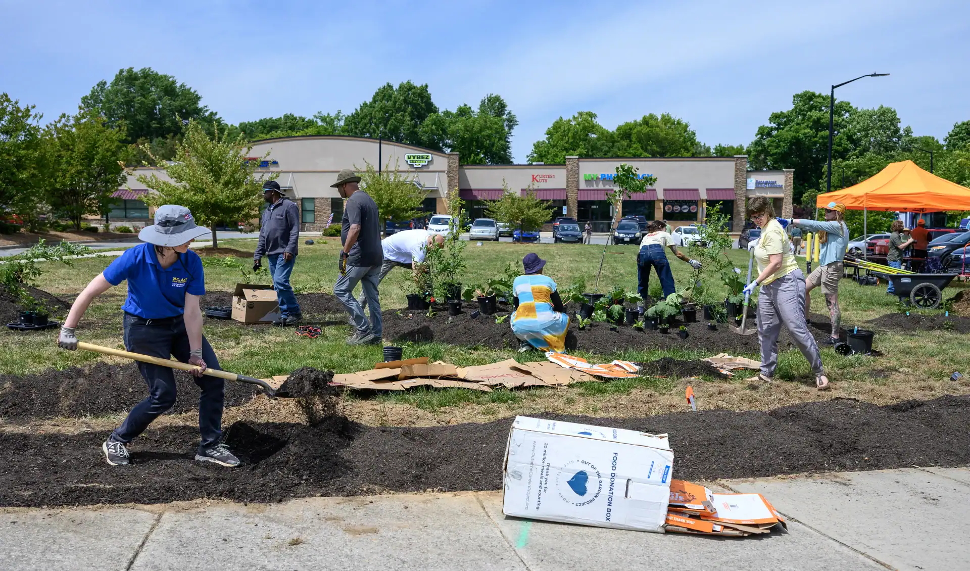 Volunteers are planting and landscaping in a grassy area near stores. Several people are using tools and arranging plants, while others work on the ground. An orange tent is visible in the background.
