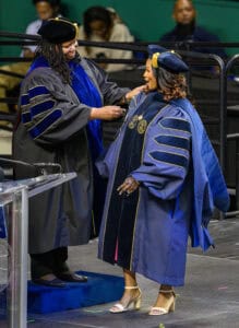 Graduation ceremony with two women in academic regalia exchanging a diploma.