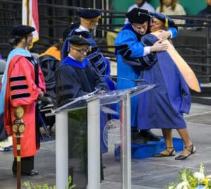 Graduates embrace during a university commencement ceremony with faculty members in academic regalia.