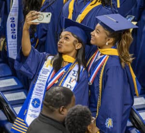 Graduates taking a selfie at graduation ceremony in blue caps and gowns with honor cords and medals.