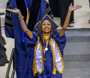 Bright smiling graduate in blue cap and gown celebrating at graduation ceremony, holding her arms up.