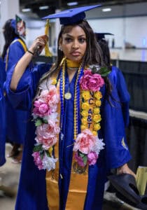 Graduation student in blue gown and cap with floral lei at ceremony.