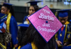 This Barbie just graduated cap with pink glitter and white text, worn by a graduate at a ceremony.