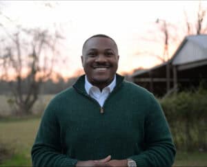 Smiling man outdoors during sunset in a green sweater and white shirt.