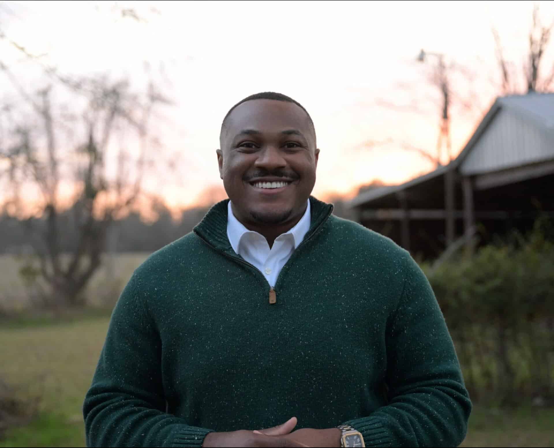 Smiling man outdoors during sunset in a green sweater and white shirt.