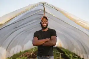 A smiling man stands with arms crossed inside a greenhouse, surrounded by rows of plants. He wears a black t-shirt and a cap, posing confidently against a backdrop of a clear sky.