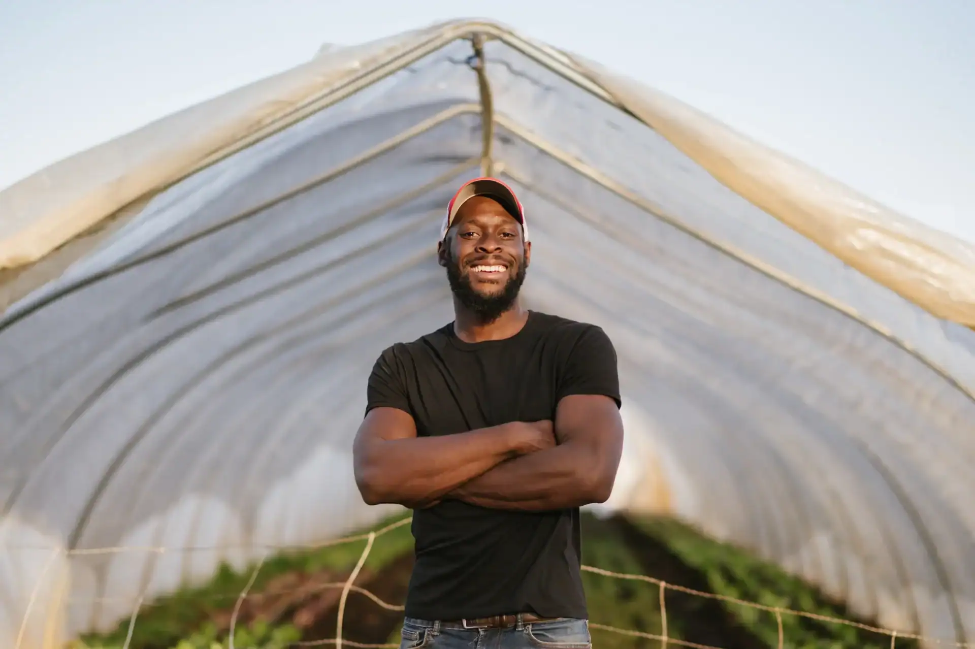 A smiling man stands with arms crossed inside a greenhouse, surrounded by rows of plants. He wears a black t-shirt and a cap, posing confidently against a backdrop of a clear sky.