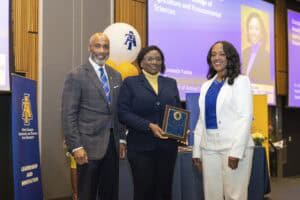 A woman receiving an award at a formal event with two colleagues, university banners, and presentati.